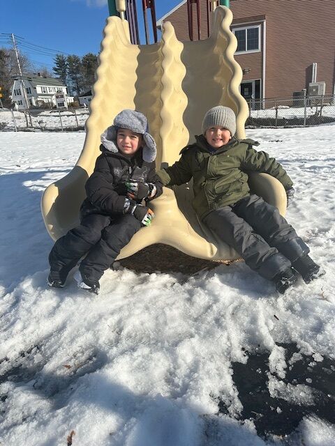 Two young boys in winter clothing sit on a playground slide covered in snow.