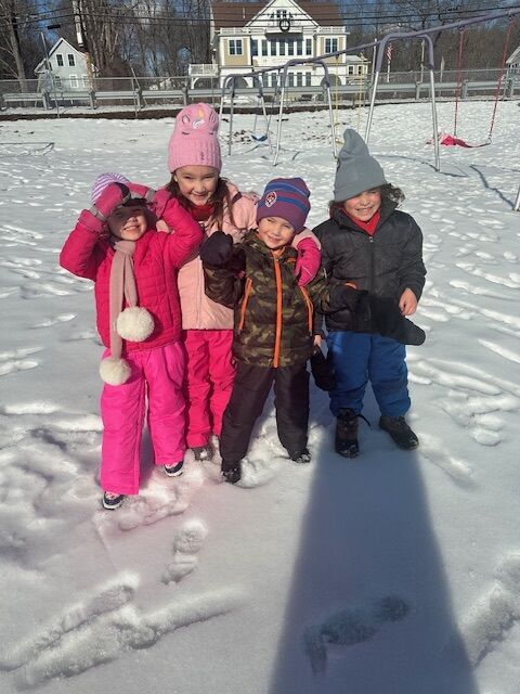 Four children in winter clothing pose in a snowy playground with swings in the background.