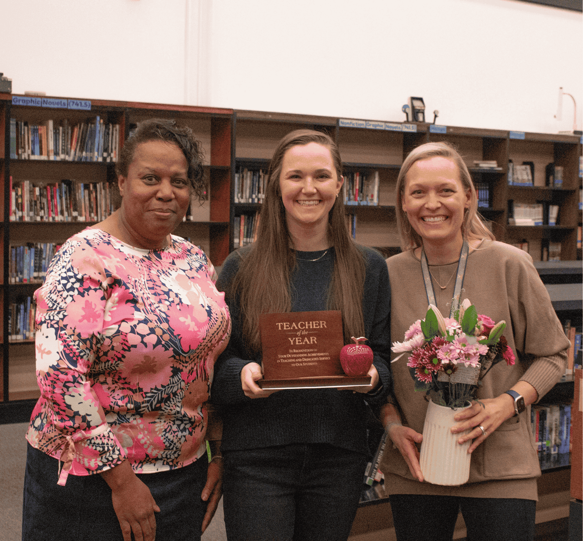 Three women stand in front of bookshelves, one holding a "Teacher of the Year" award and a red apple.