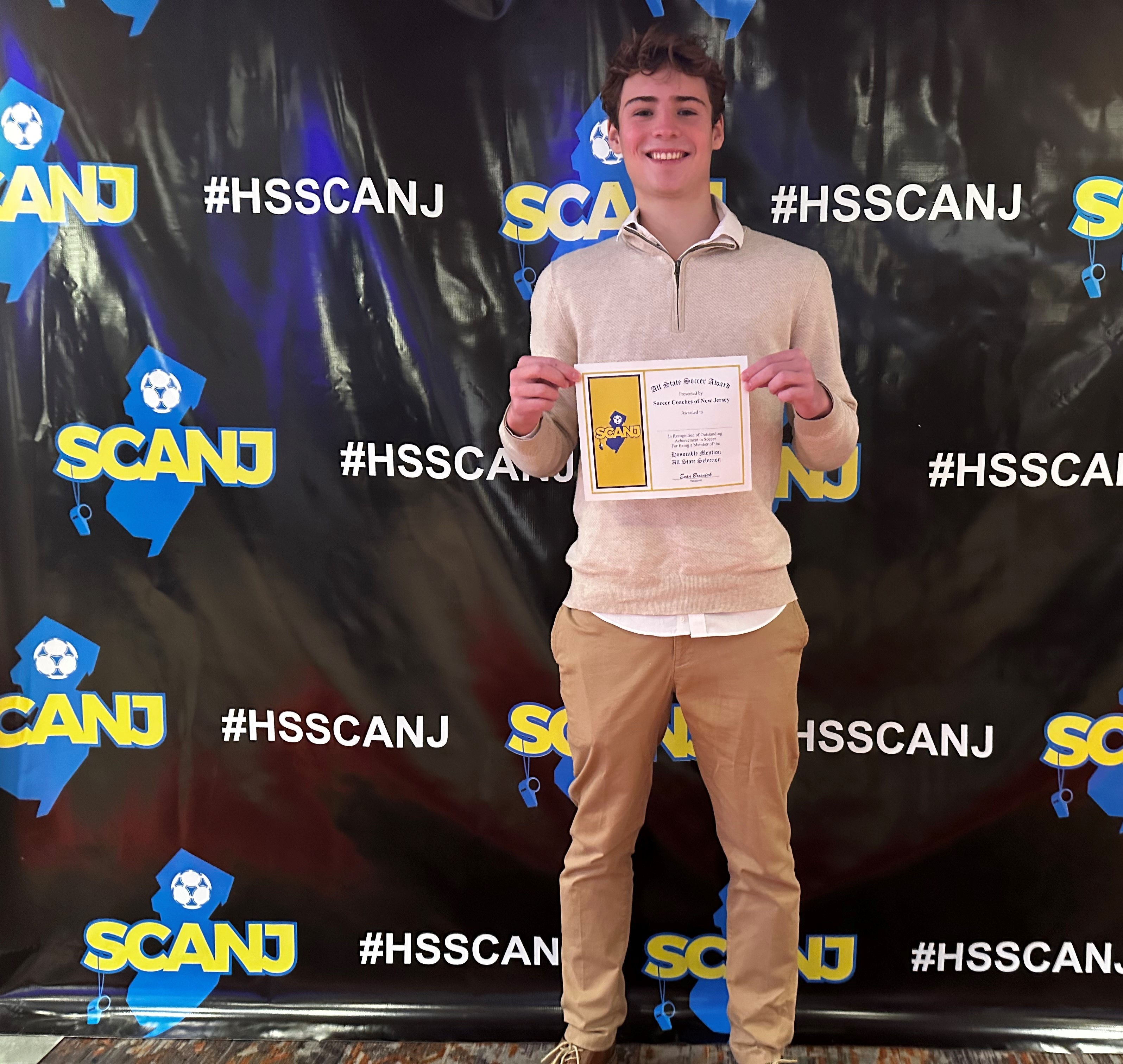 A smiling young man holds an 'All State Soccer Award' certificate in front of a backdrop with 'SCANJ' and '#HSSCANJ' logos.