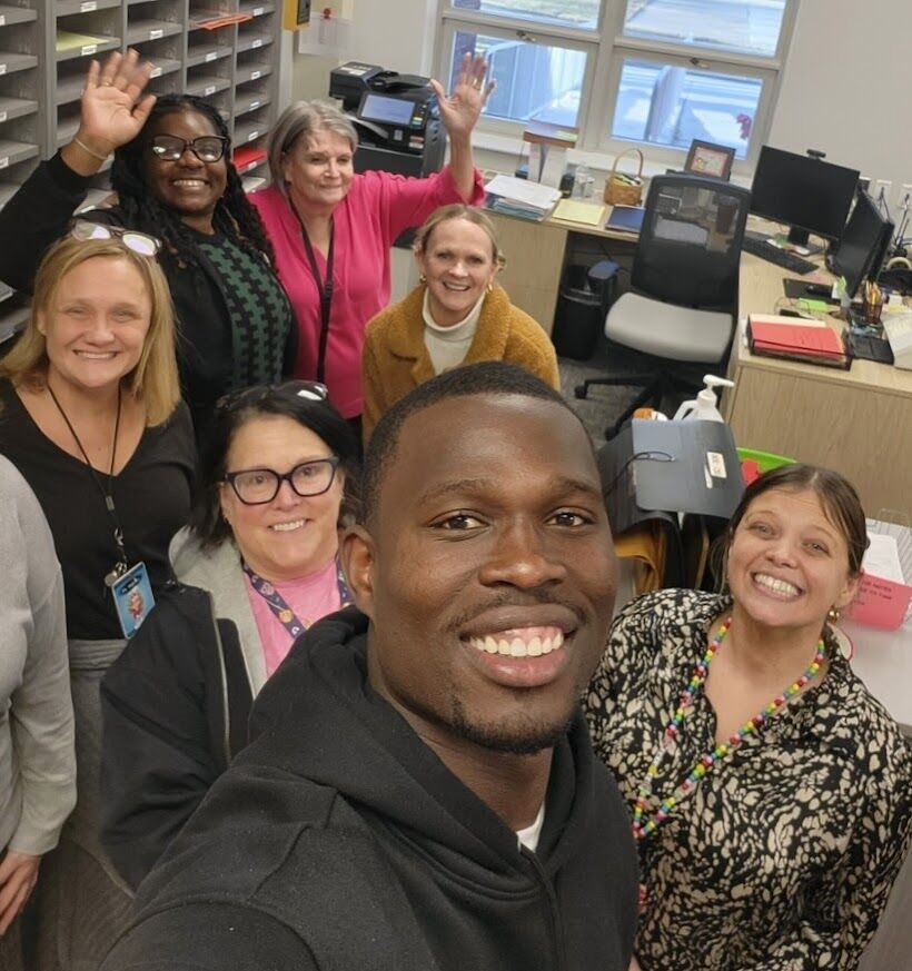A diverse group of smiling colleagues wave hello from an office setting.