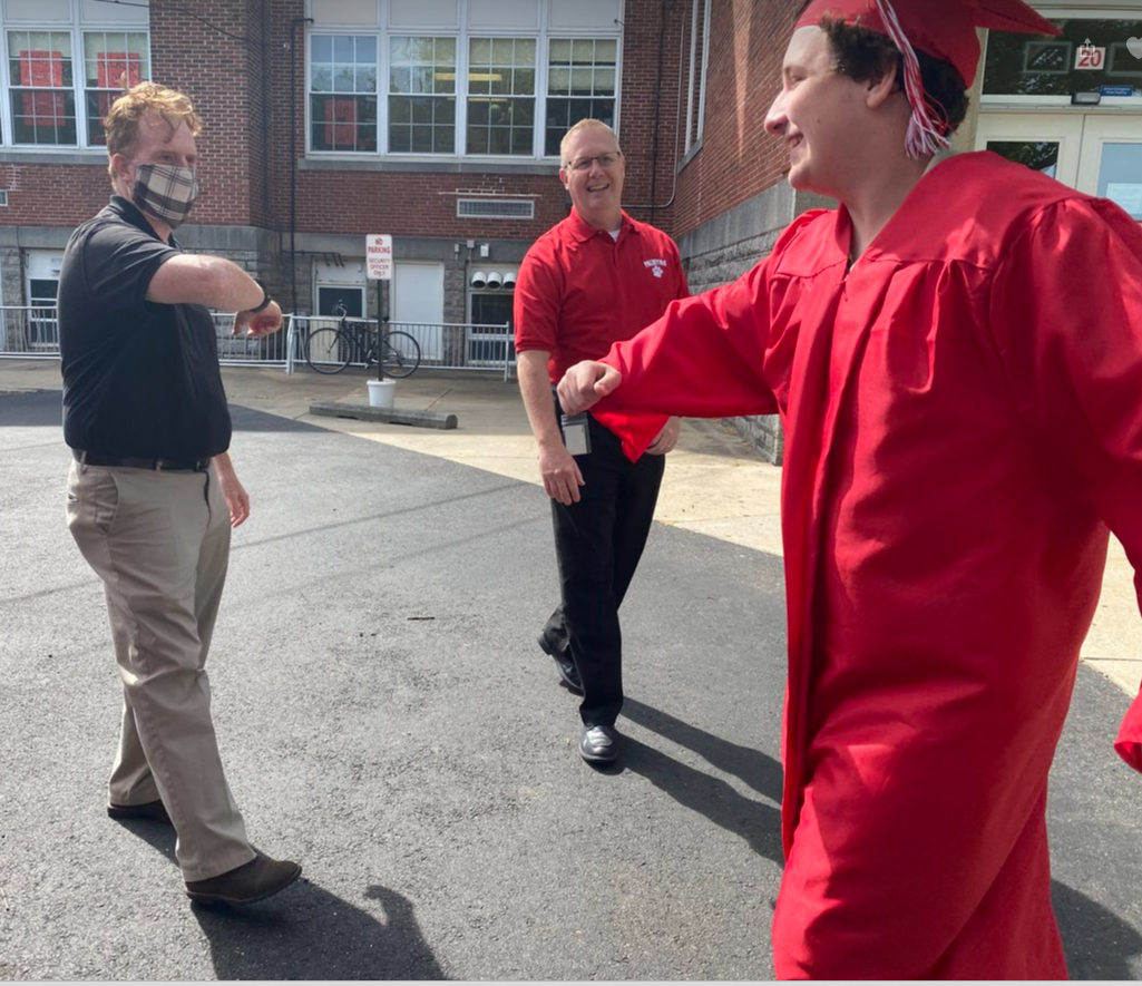 A graduate in a red cap and gown bumps elbows with a masked man, while another man in a red shirt smiles.