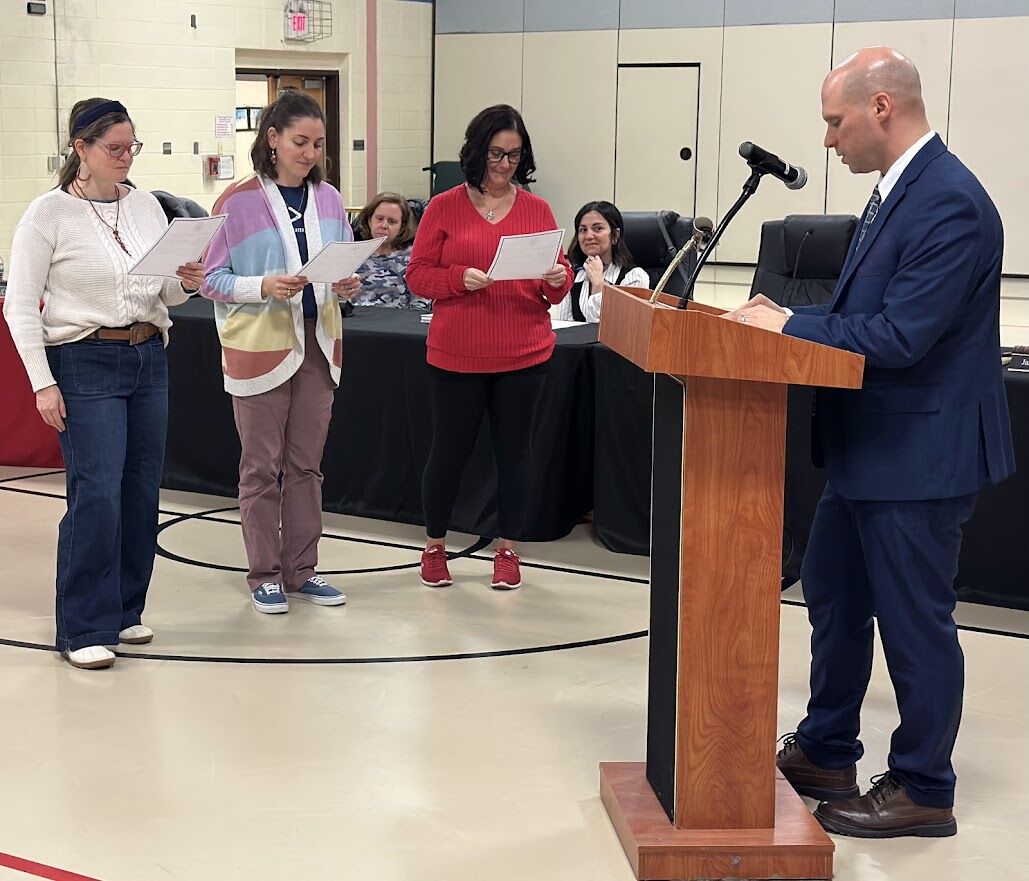 A man in a suit stands at a podium, addressing three women holding papers.