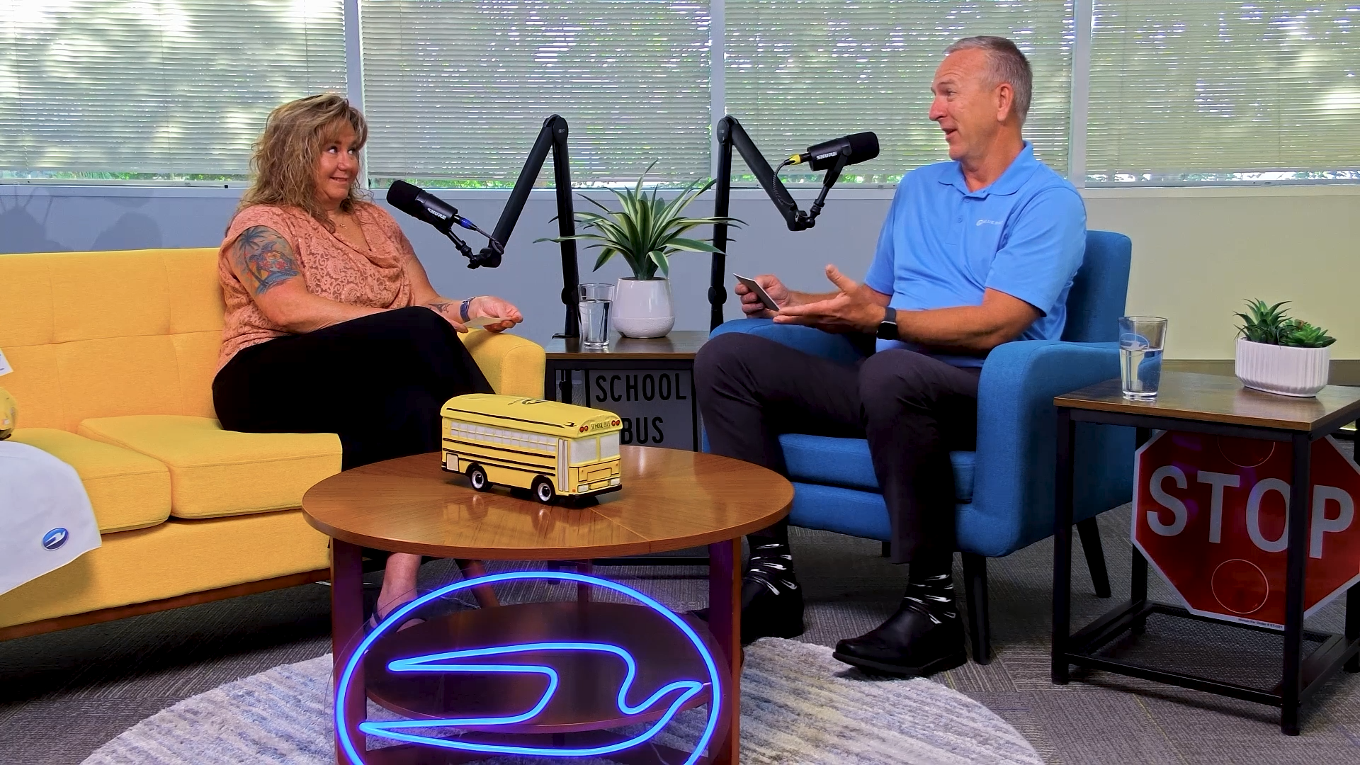 Two people sit in chairs and on a couch, facing each other with microphones and a school bus model on a table between them.