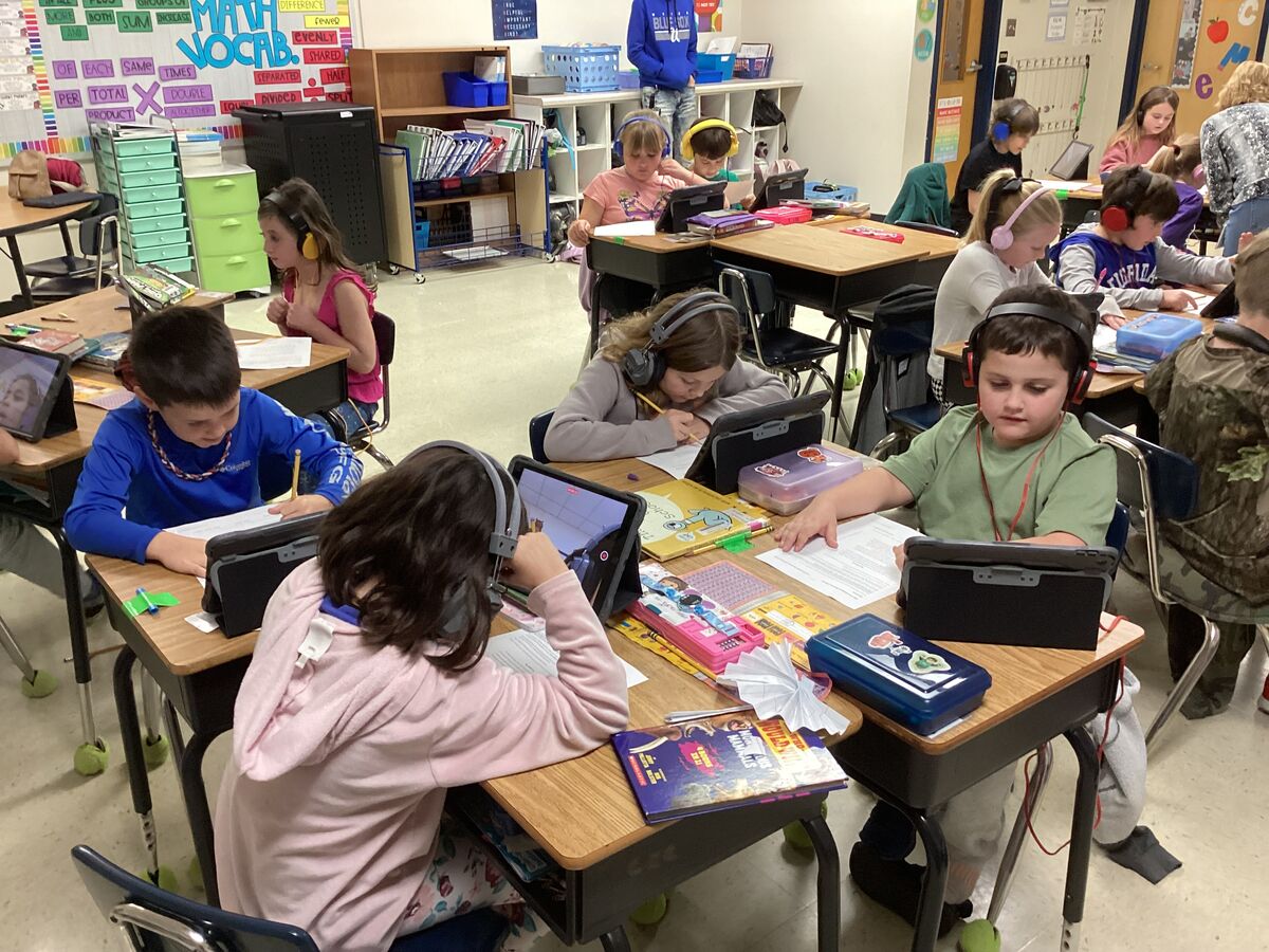 Students wearing headphones work at desks with tablets and papers in a classroom.