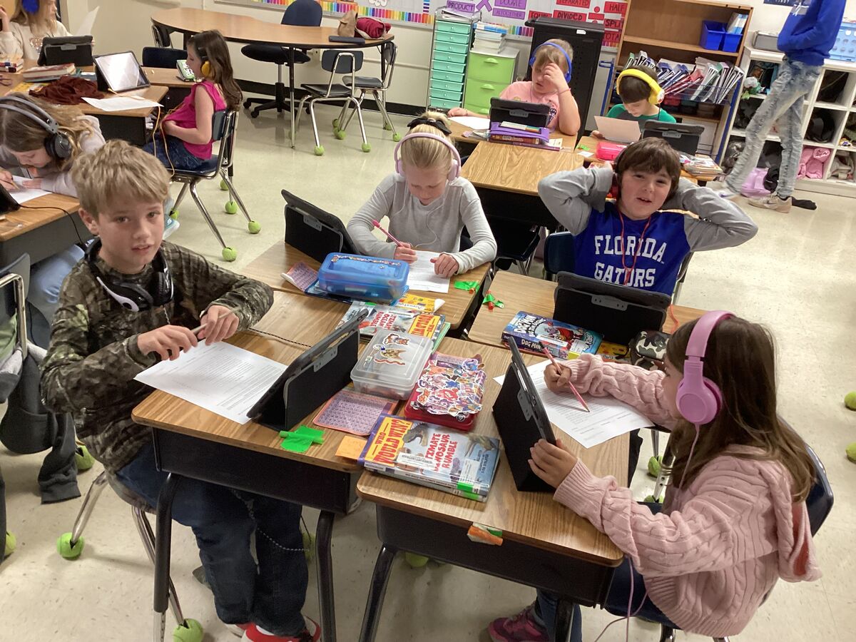Students wearing headphones work at desks with tablets and papers in a classroom.