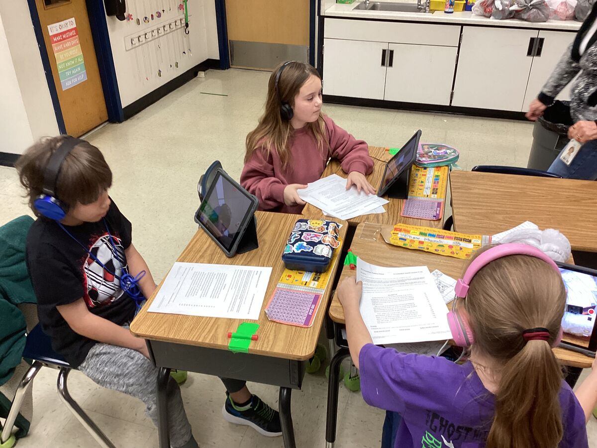 Three students wearing headphones work at desks with tablets and papers in a classroom.