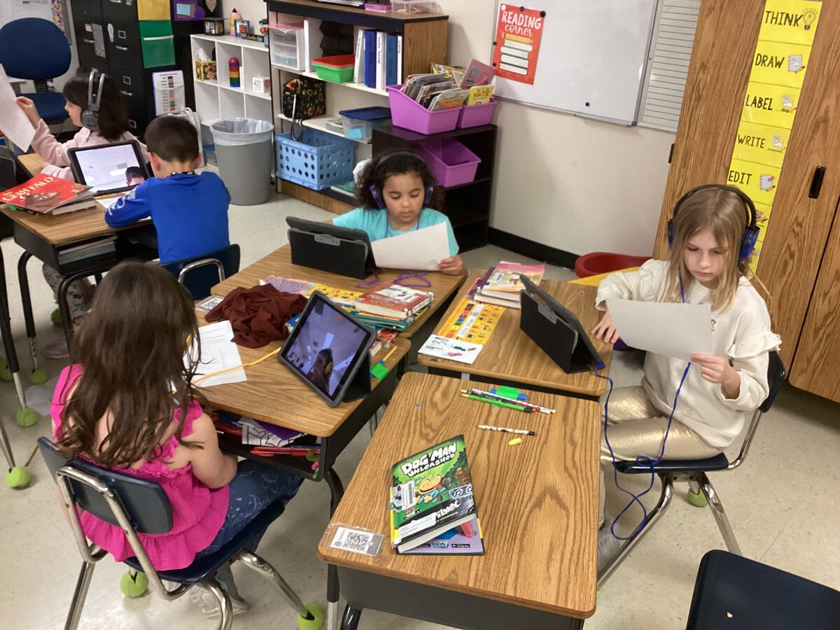 Students wearing headphones work at their desks with tablets and papers in a classroom.