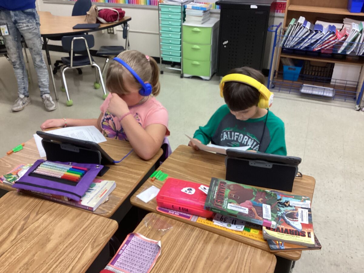 Two children wearing headphones focus on their tablets and books at individual desks in a classroom.