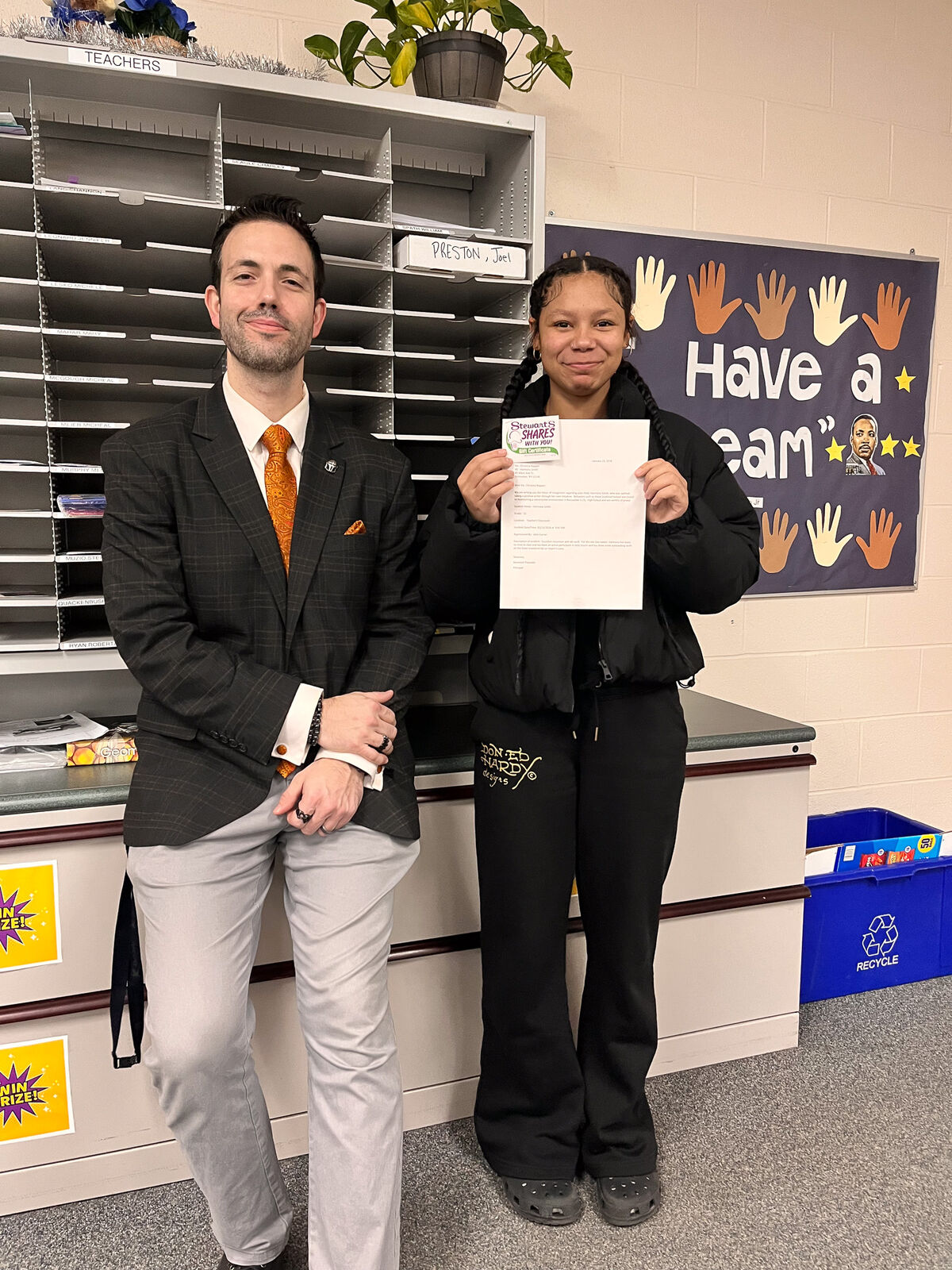A man in a suit and a young woman stand together, holding a gift certificate.