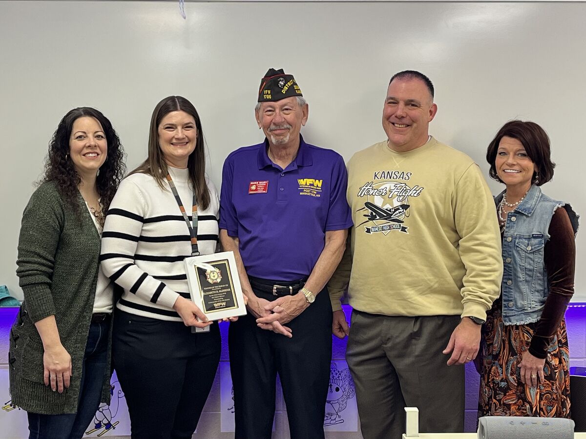 Five people pose indoors; a woman holds an award plaque next to an older man in a VFW cap and purple shirt, with three others including a man in a “Kansas Honor Flight” sweatshirt, all standing in front of a classroom whiteboard with purple LED lighting.