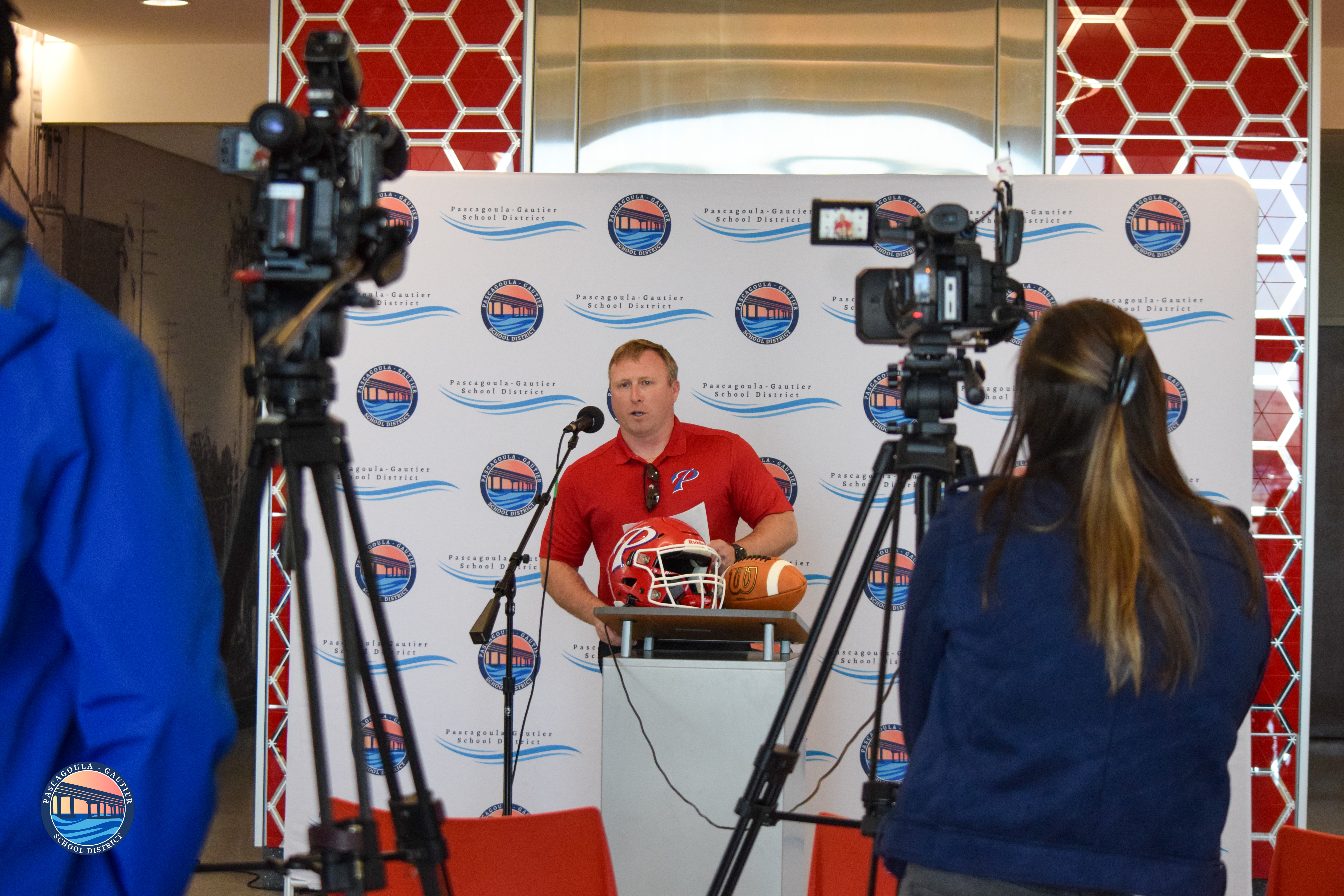 A man in a red polo shirt speaks at a podium with a football helmet and ball, facing two cameras.