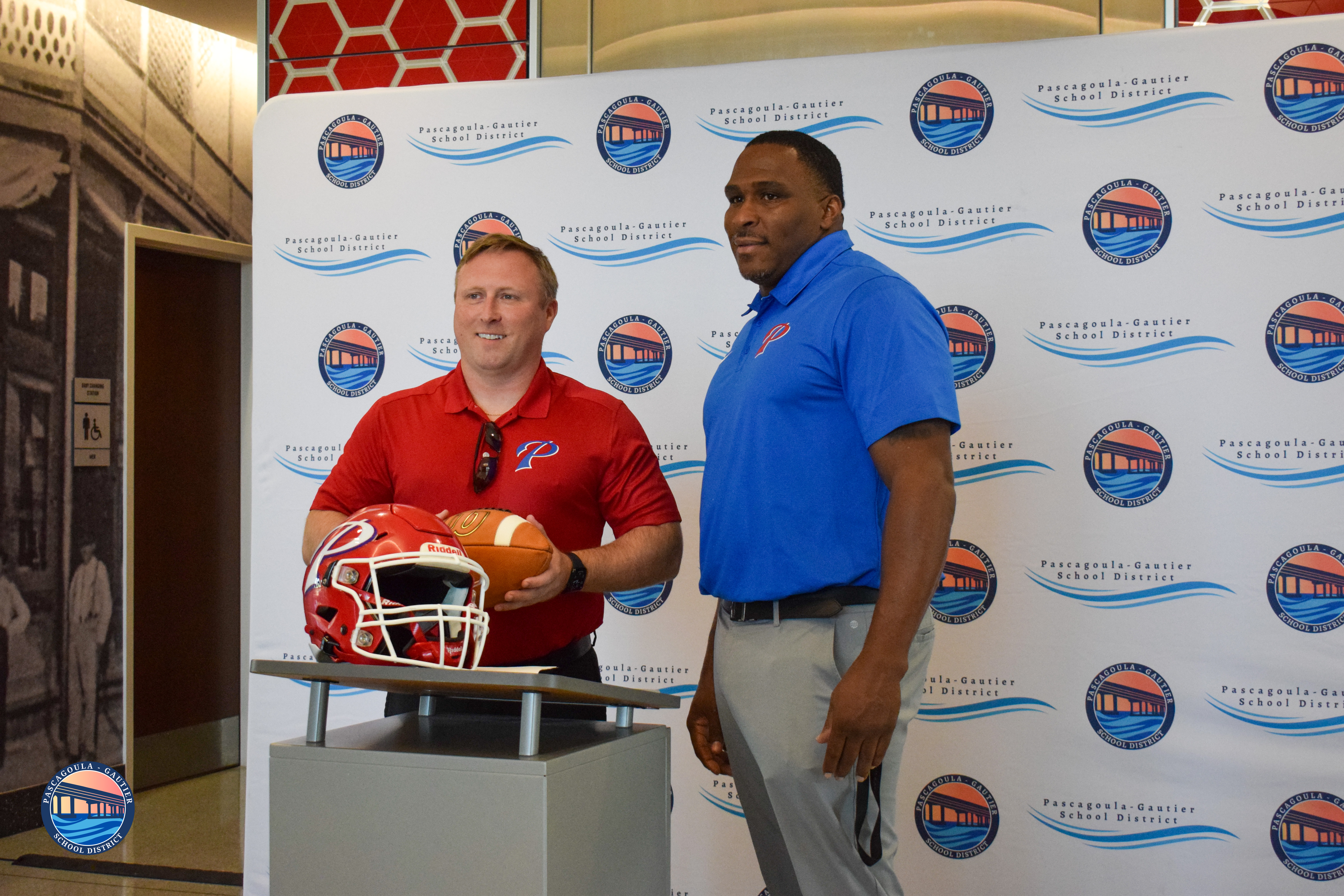 Two men stand in front of a banner with the Pascagoula-Gautier School District logo. The man on the left wears a red shirt and holds a football helmet and football.