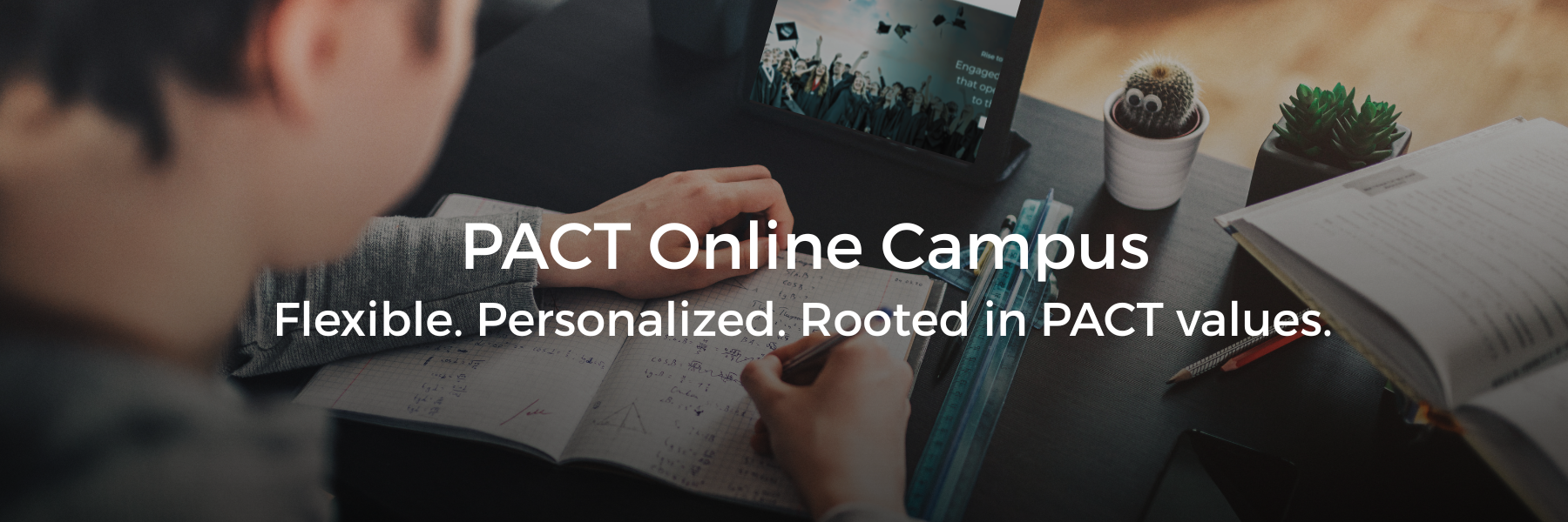 A person's hands write in a notebook on a desk with a tablet displaying graduates.