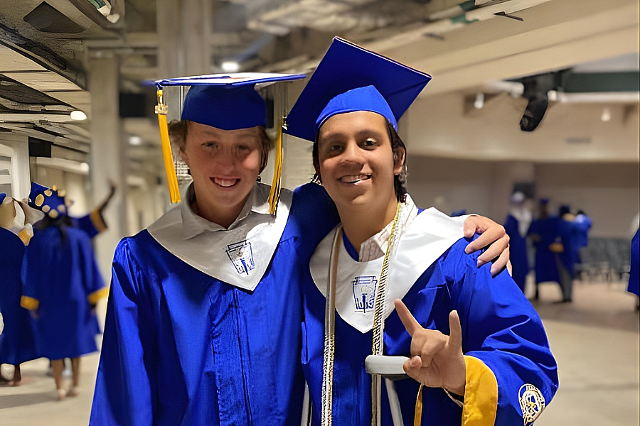 Two smiling graduates in blue caps and gowns pose together, one with an arm around the other.