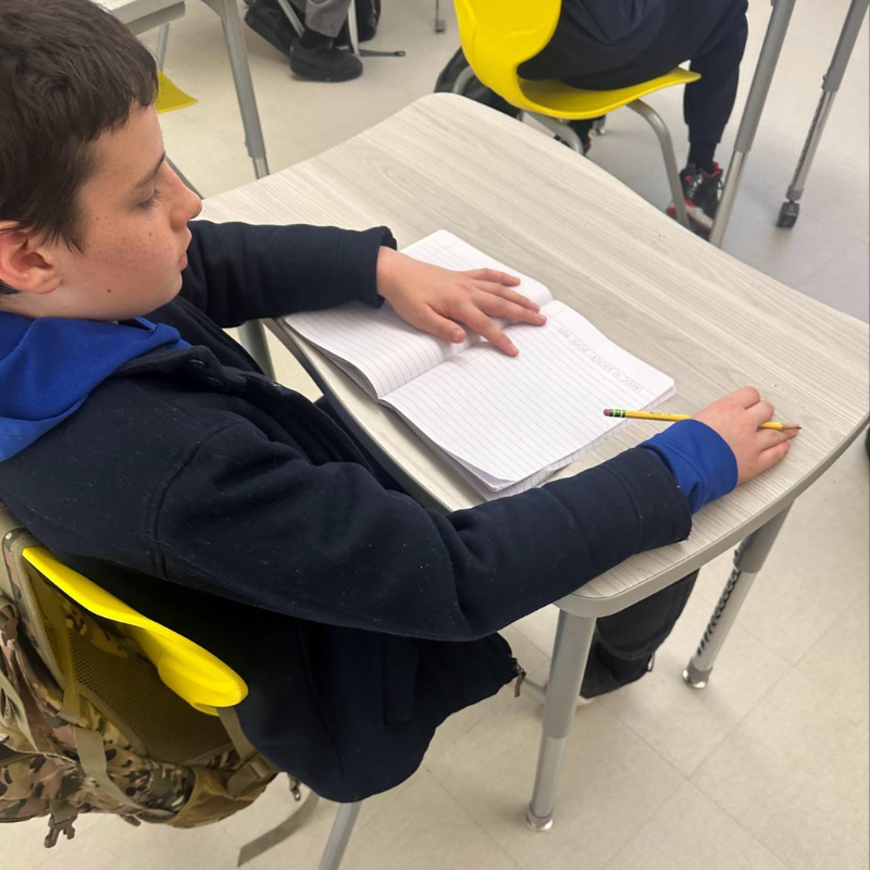 A young student sits at a desk, looking down at an open notebook with a pencil in hand.
