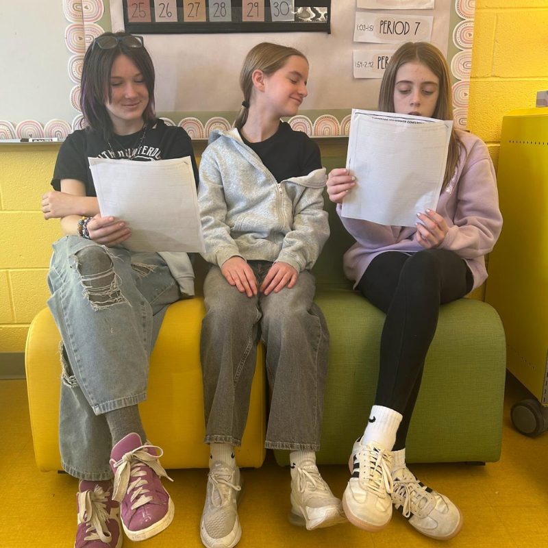 Three students sit on a colorful couch in a classroom, each holding a piece of paper.