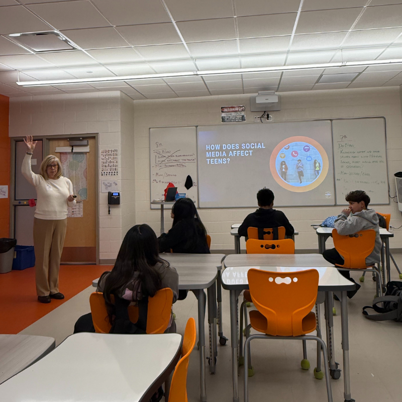 A teacher gestures while presenting a slide titled "HOW DOES SOCIAL MEDIA AFFECT TEENS?" to students in a classroom.