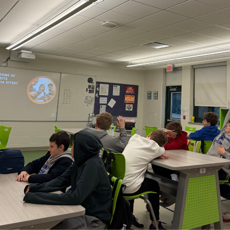 Students sit at desks in a classroom, facing a projector screen displaying text and images.