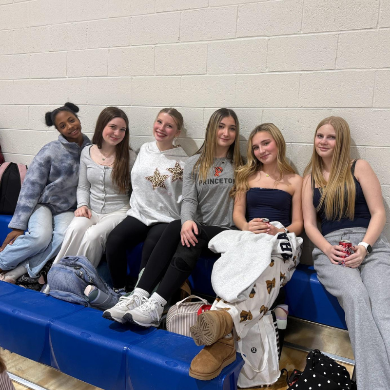 Six young women sit together on a blue bench against a brick wall.