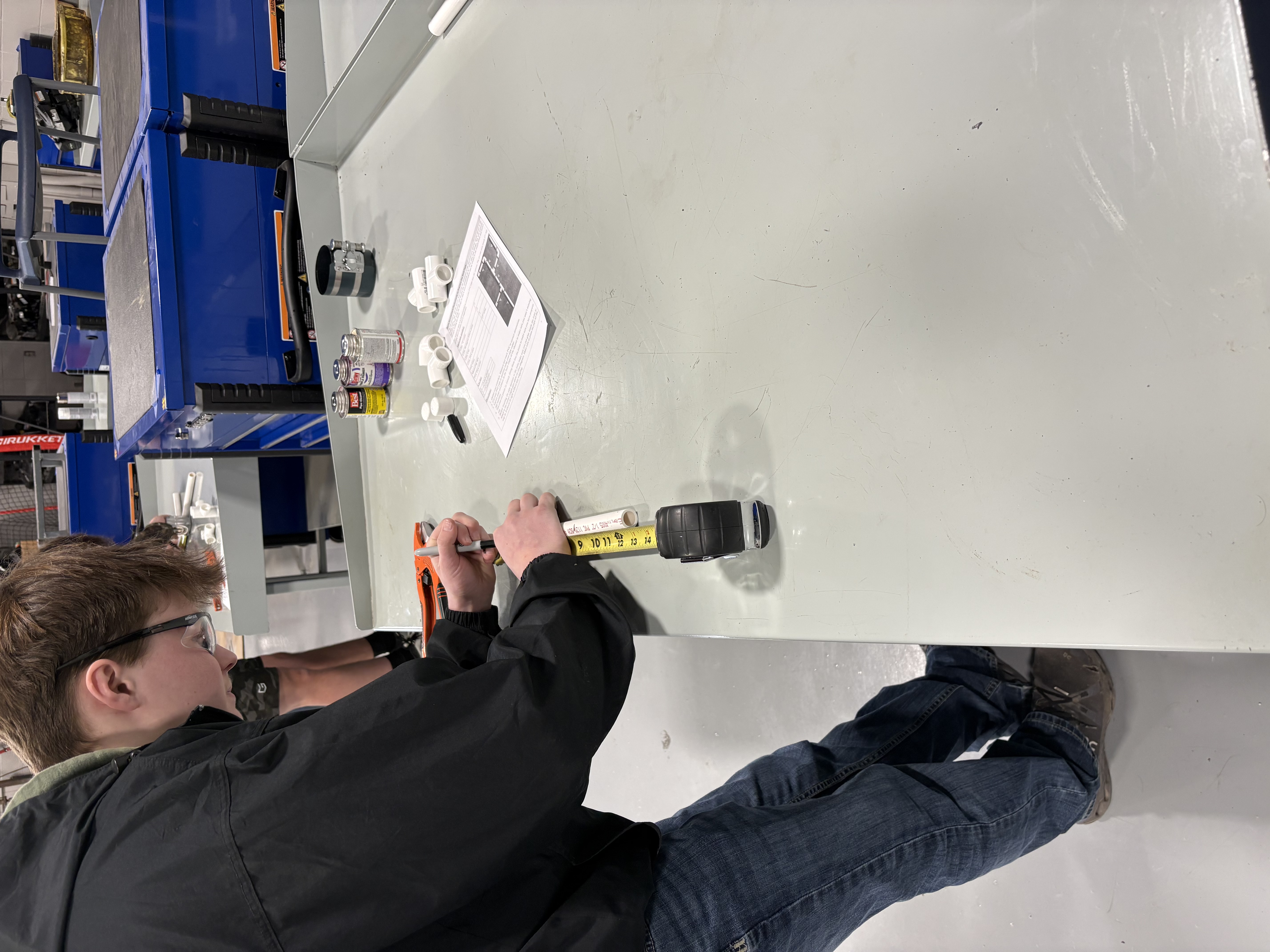 A student measures a piece of pipe with a tape measure and marker.