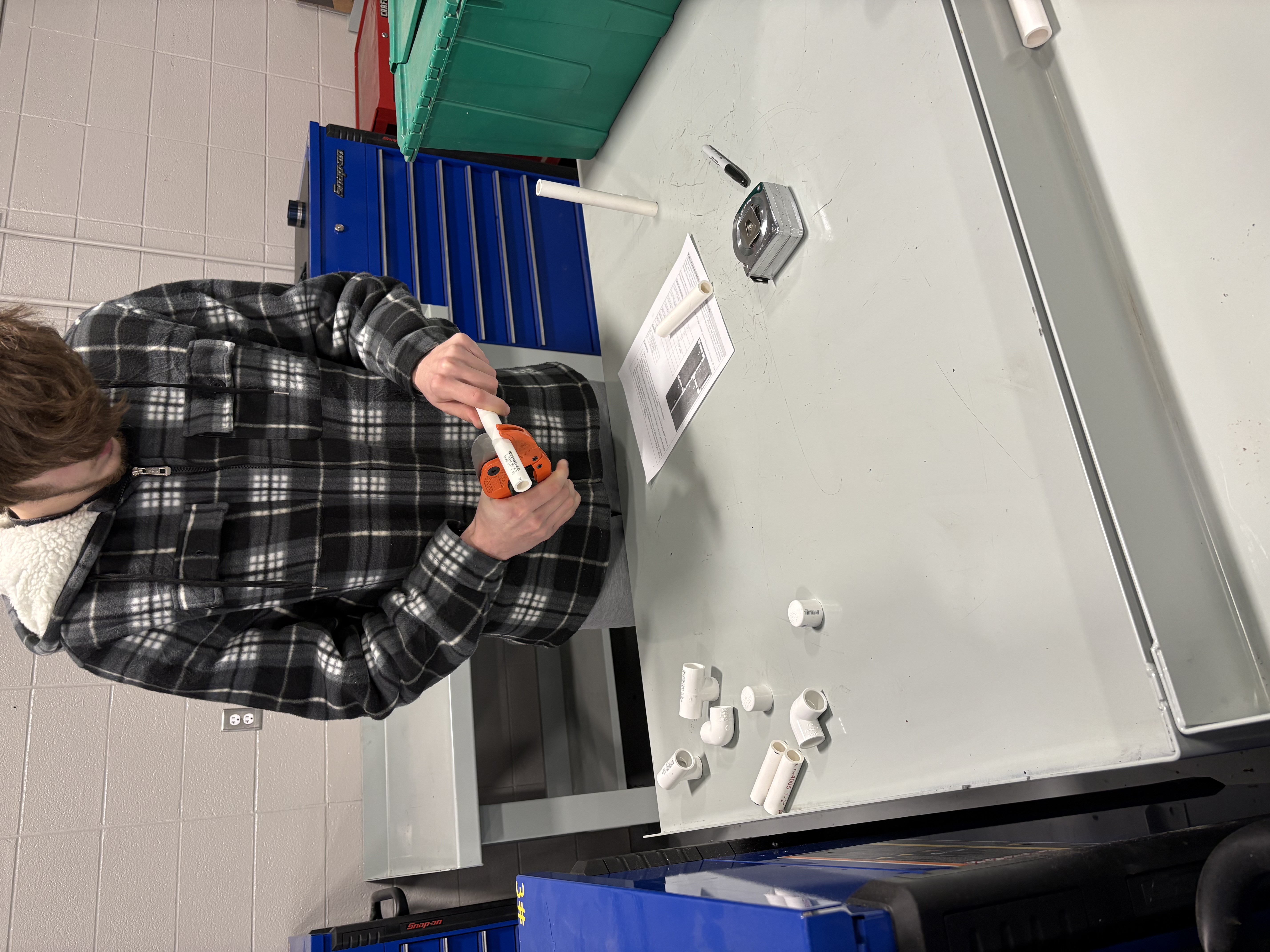 Student uses a pipe cutter on a white PVC pipe at a workbench.