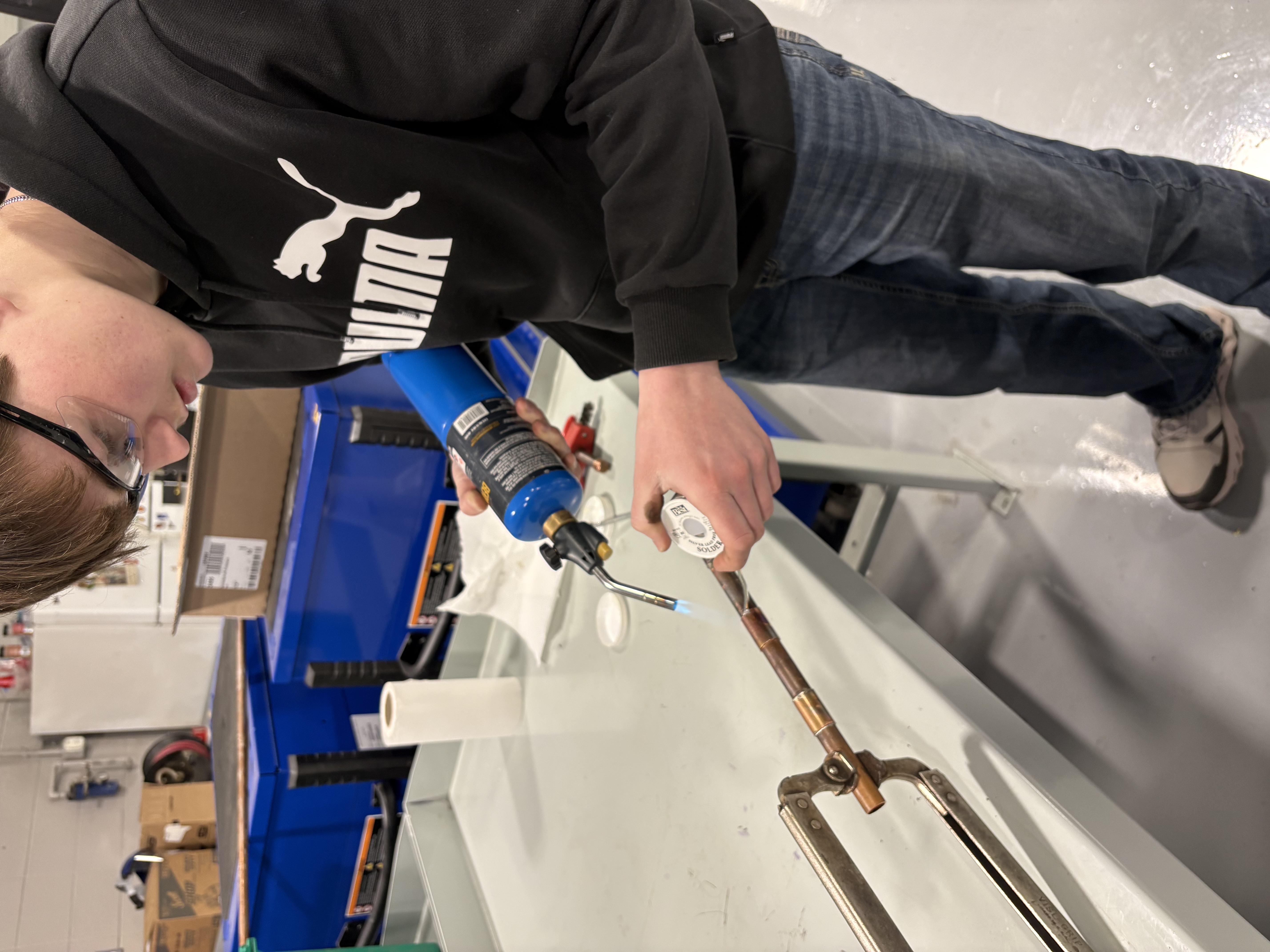A student uses a propane torch and solder to work on a copper pipe.