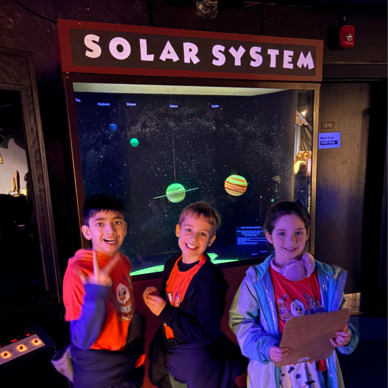 Three children smile in front of a "SOLAR SYSTEM" exhibit displaying planets.