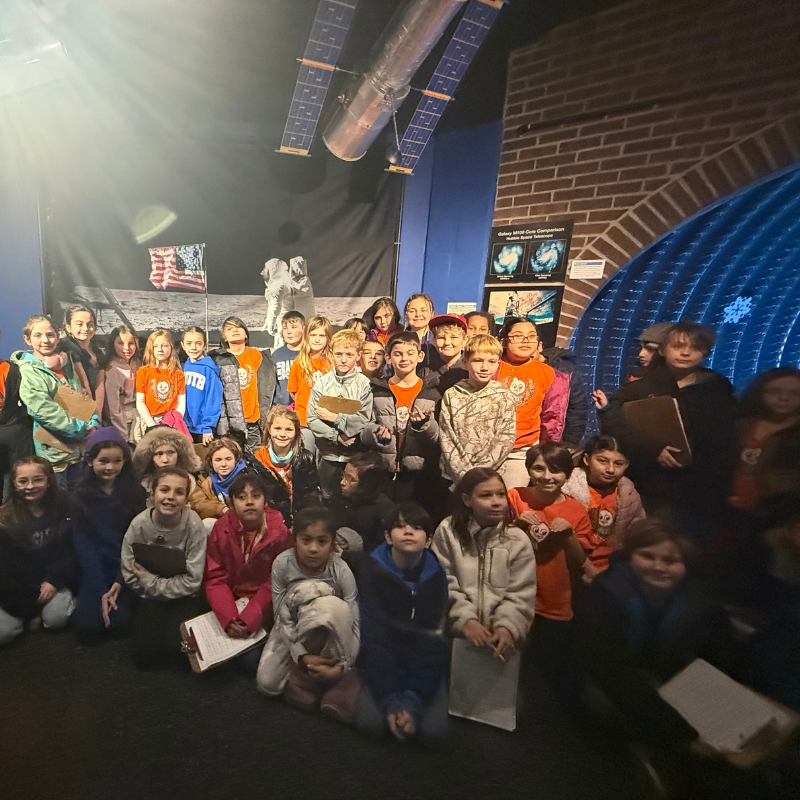 A group of children pose for a photo in a museum exhibit featuring a space station model and moon landing diorama.