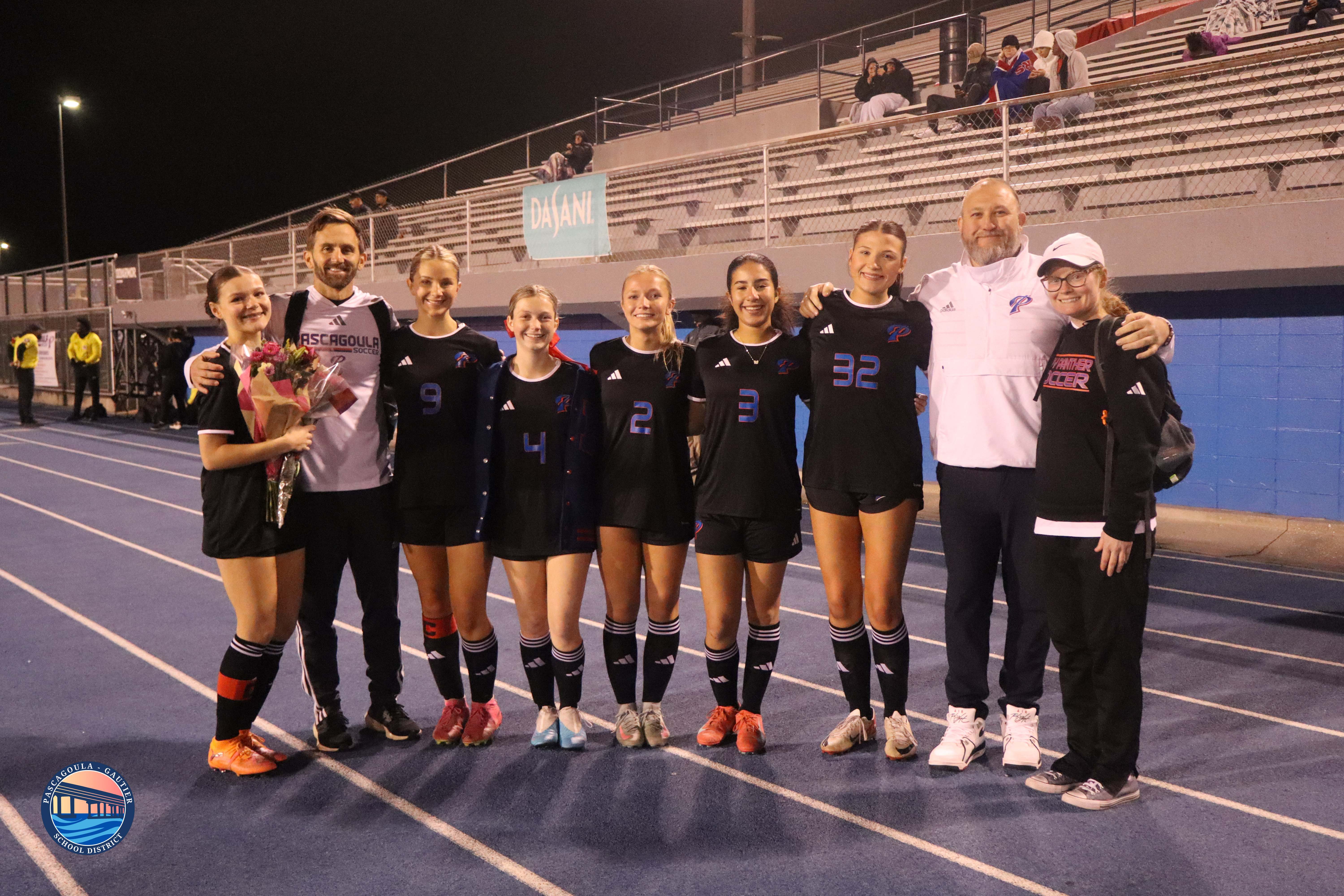 A soccer team poses for a photo on a blue track at night, with coaches and players smiling.