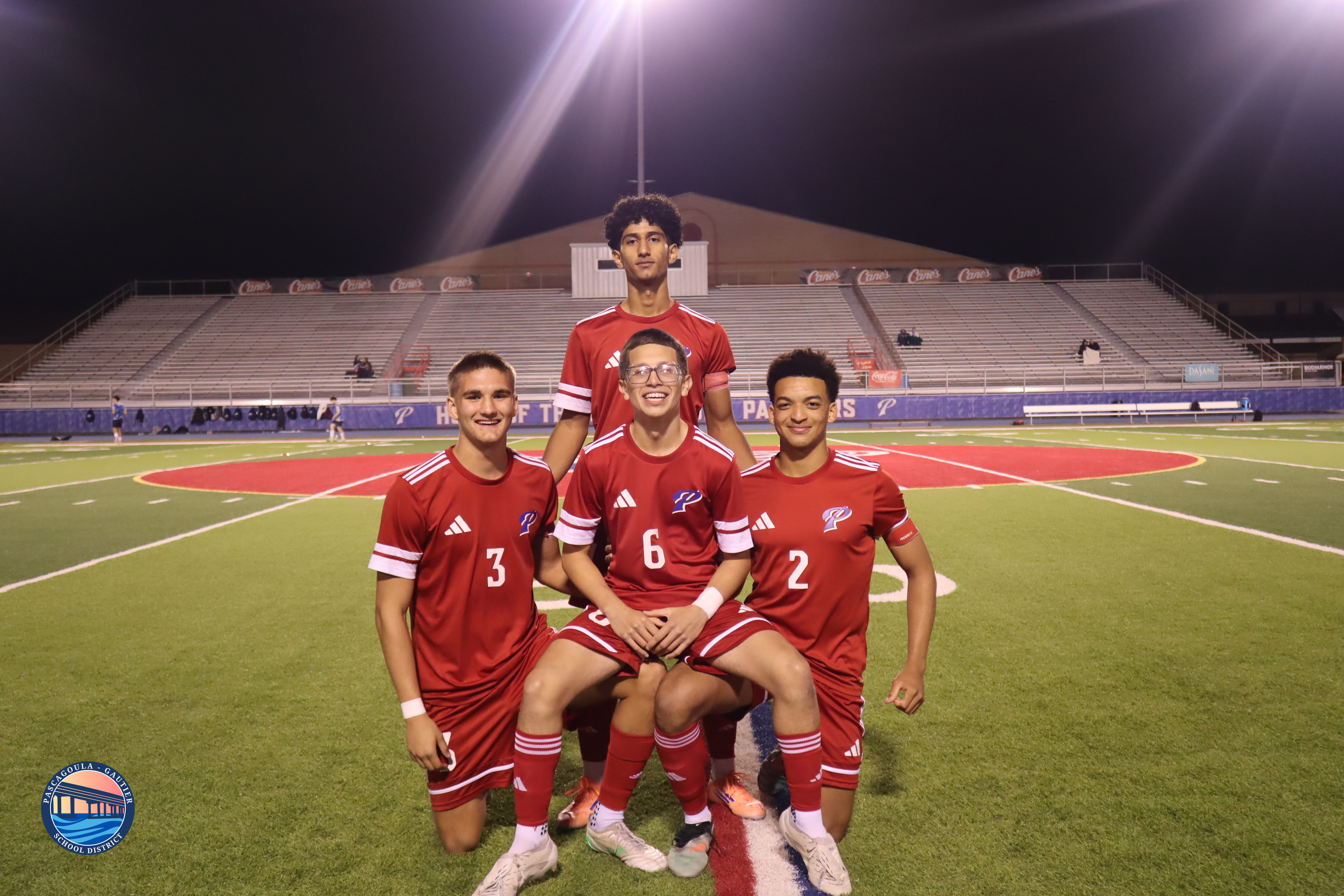 Four young men in red soccer uniforms pose on a green field at night, with stadium seating in the background.
