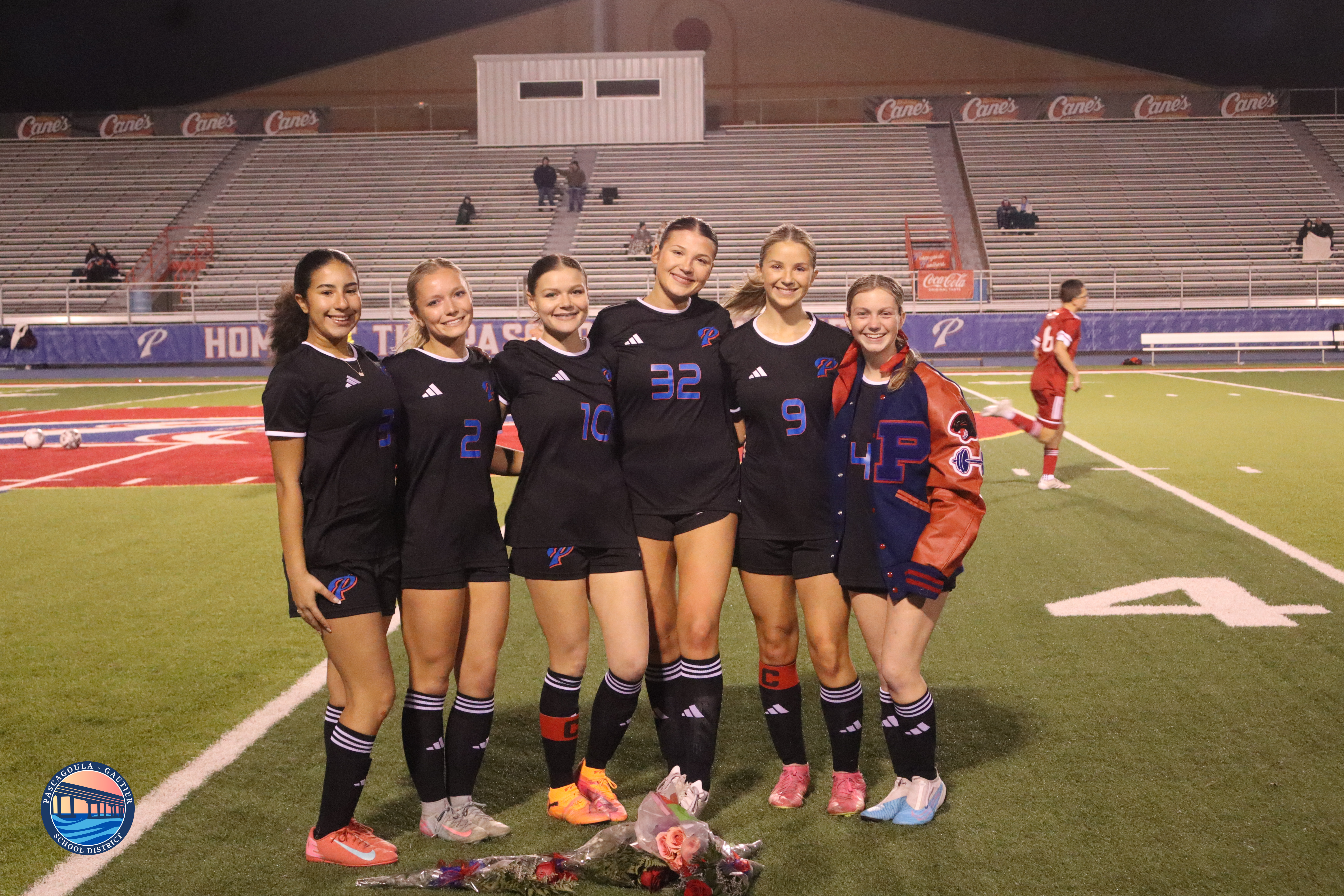 Six smiling young women in soccer uniforms pose on a field at night, with stadium seating in the background.