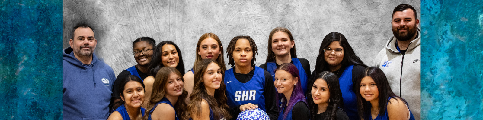 A basketball team poses for a photo with their coaches against a textured gray backdrop.
