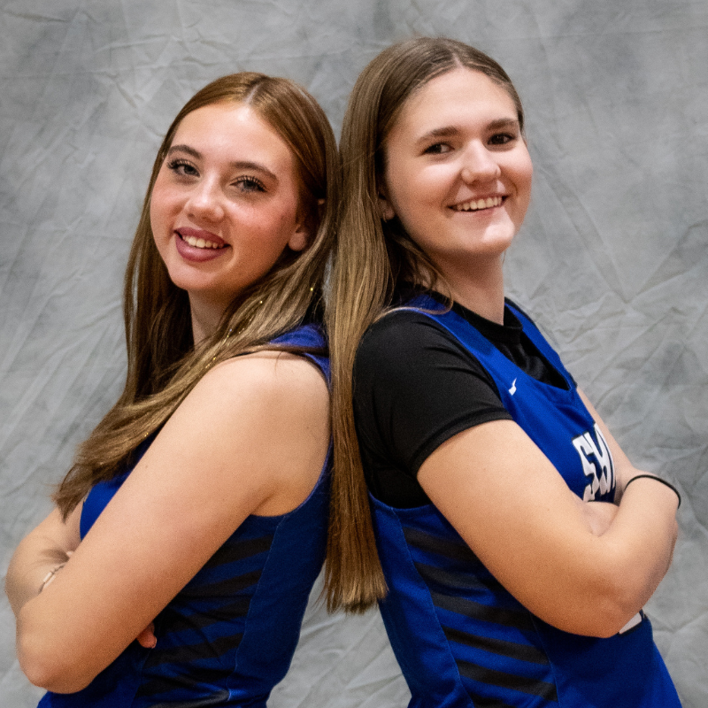 Two young women in blue athletic jerseys stand back-to-back with arms crossed.