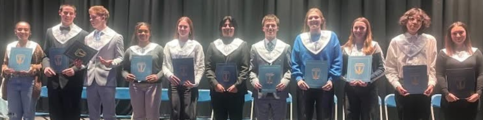 A group of students stand on a stage, holding certificates and wearing academic regalia.
