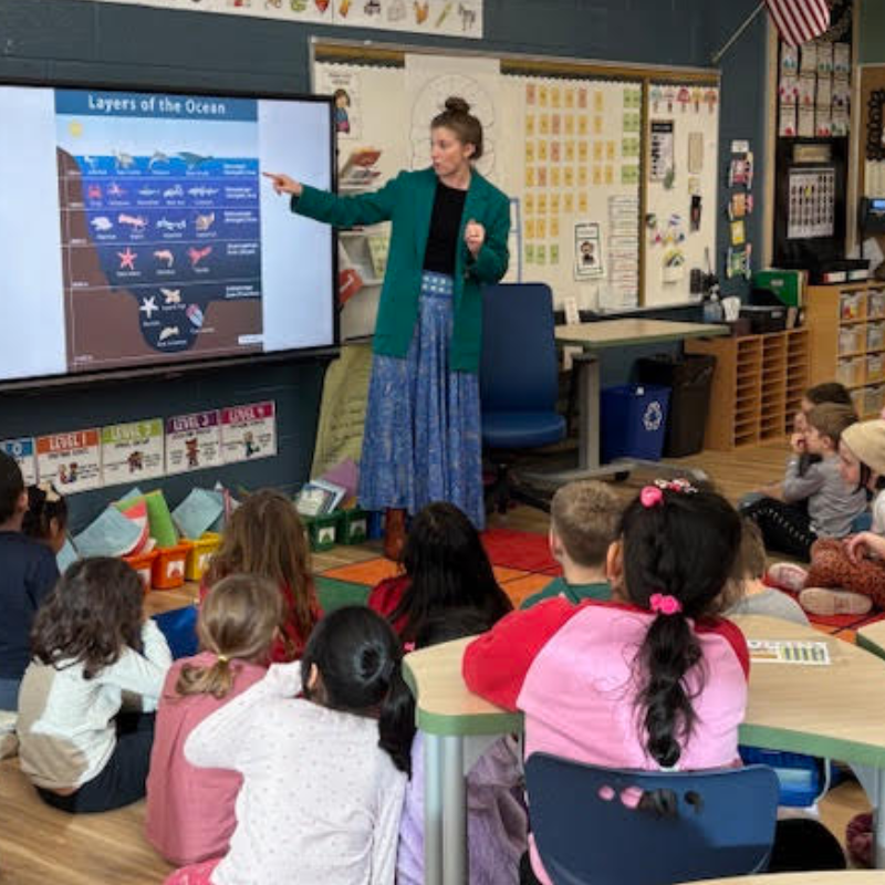 A teacher points to a diagram of the 'Layers of the Ocean' on a large screen to a class of young students.