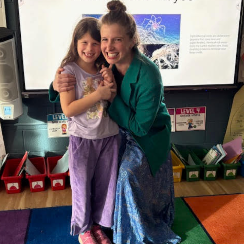 A woman in a green jacket hugs a young girl in purple pajamas in front of a large screen.