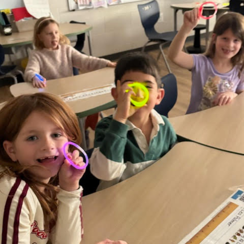 Children in a classroom hold up colorful glow bracelets, smiling and playing.