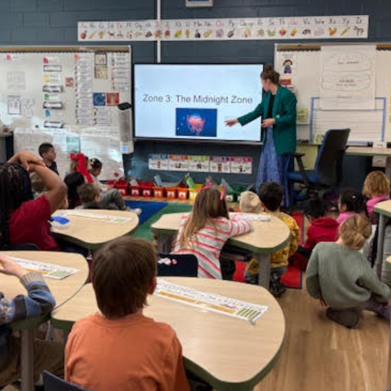 A teacher points to a screen displaying "Zone 3: The Midnight Zone" to a class of young students.