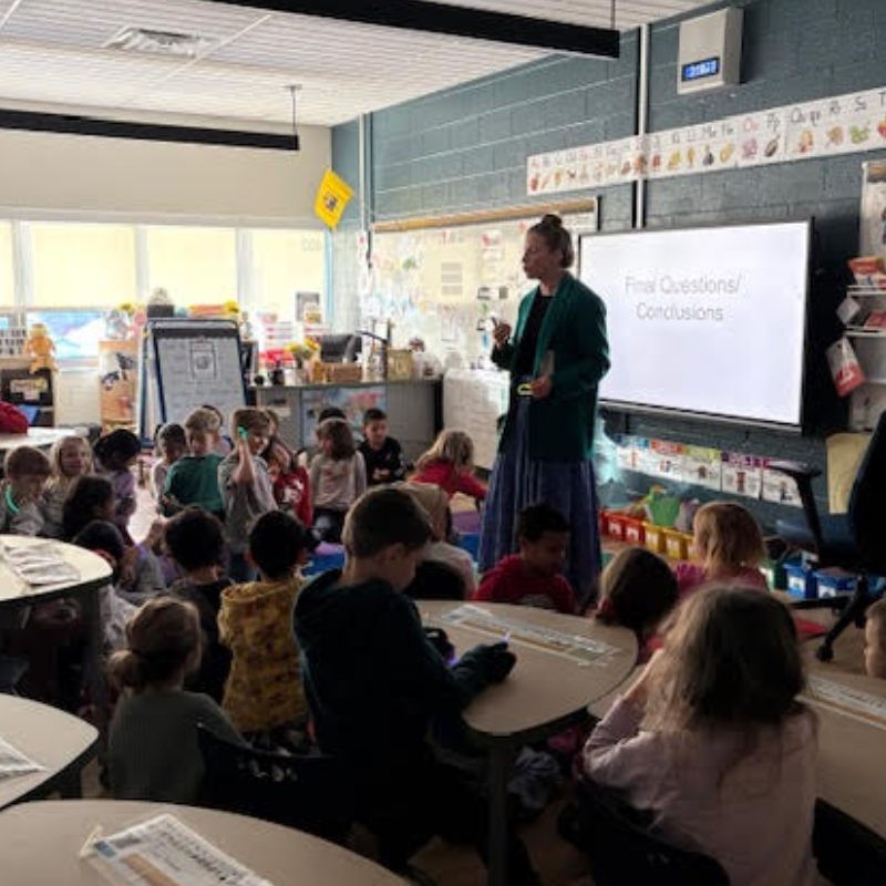 A teacher stands before a class of young students, presenting on a screen that reads 'Final Questions/Conclusions'.