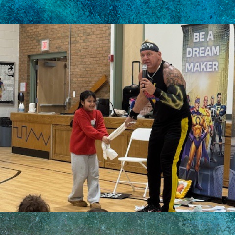 A man in athletic wear holds a microphone and interacts with a young girl in a red sweater, both pulling on a white cloth.