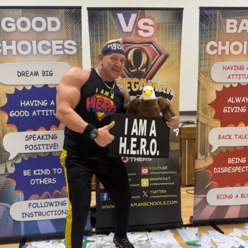 A muscular man in a 'I AM A HERO' shirt holds a stuffed eagle and a sign that reads 'I AM A H.E.R.O.'