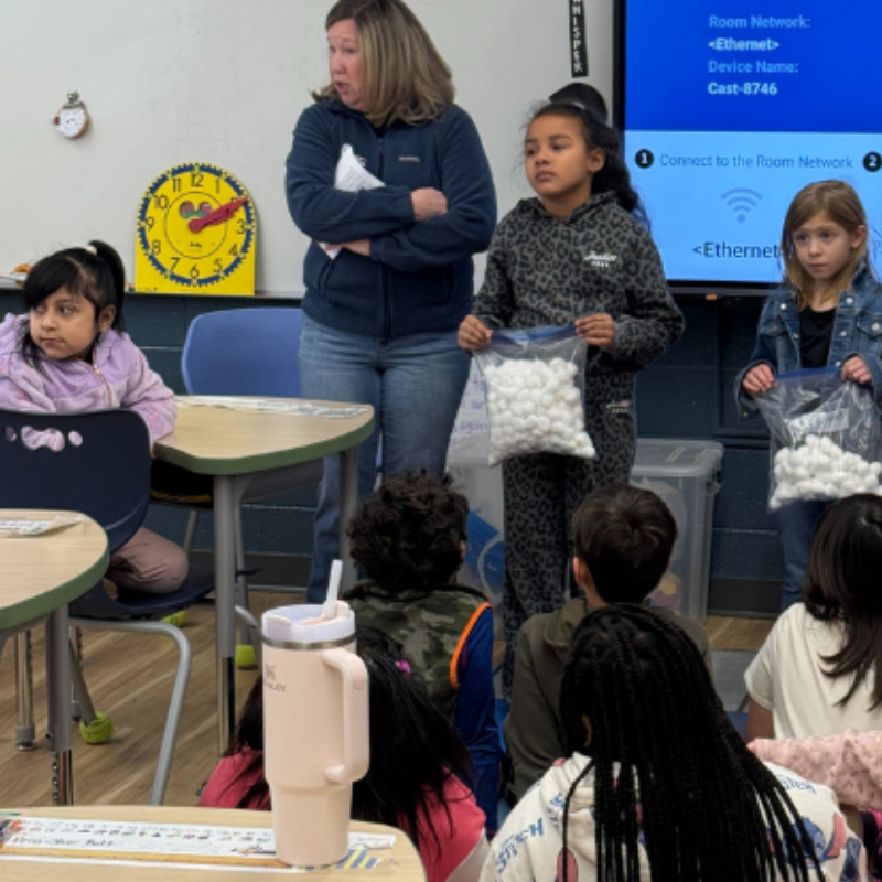 A teacher stands in front of a classroom of students, some holding bags of white pom-poms.