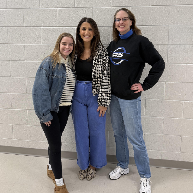 Three women stand together in front of a white brick wall, smiling.