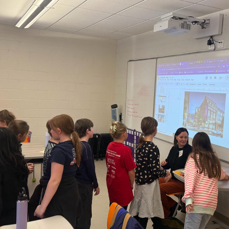 A teacher presents an image of a building on a projector screen to a group of students in a classroom.