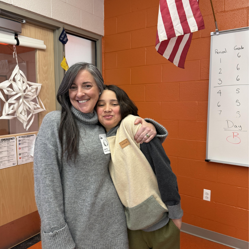 A woman and a child embrace warmly in a classroom setting with an American flag and whiteboard visible.