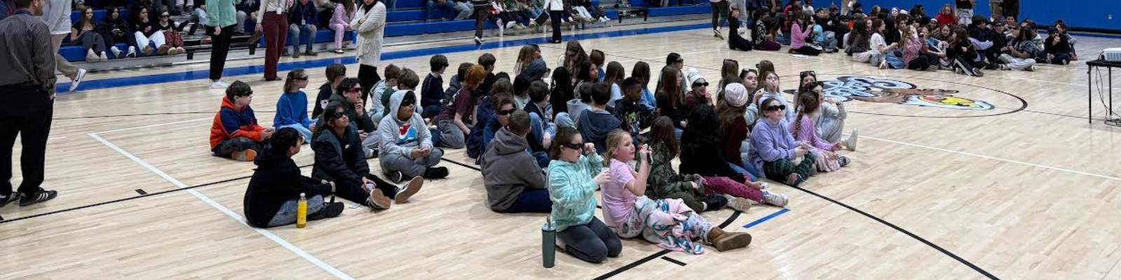 Students wearing 3D glasses sit on a gymnasium floor, watching a presentation.