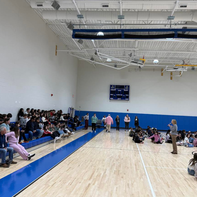 A group of students sits on bleachers and the floor of a gymnasium, listening to speakers.