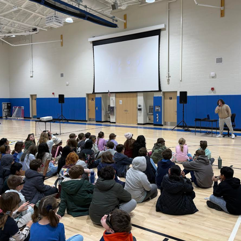 A group of children sit on a gymnasium floor watching a presentation on a large screen.