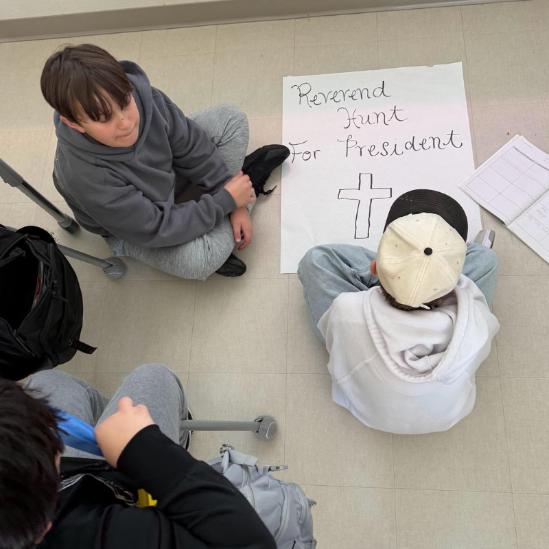 Three young people sit on the floor around a poster that reads "Reverend Hunt For President" with a drawing of a cross.